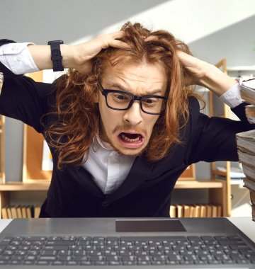 Eine Person mit langen Haaren und Brille sieht gestresst aus, während sie an einem Schreibtisch mit großen Stapeln von Papierkram und einem Laptop sitzt.