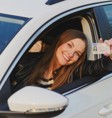 Junge Frau sitzt am Steuer und zeigt fröhlich ihren Führerschein aus dem Fenster