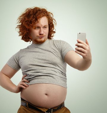 Mann mit Bierbauch in bauchfreiem Oberteil vor Smartphone
