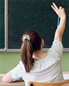 Eine Schülerin, die an einem Schreibtisch sitzt, hebt ihre Hand vor einer Tafel in einem Klassenzimmer.