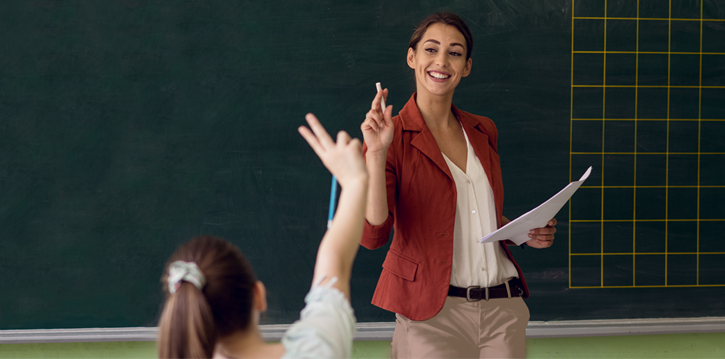 Ein Lehrer steht lächelnd mit Kreide vor einer Tafel, während ein Schüler eine Hand hebt, um eine Frage zu beantworten.